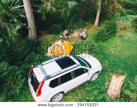 Couple Sitting Near Campfire Drinking Tea. Yellow Tent And White Suv Car. Active Leisure Time