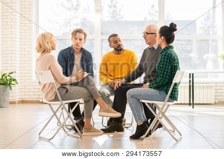 Several young and mature intercultural groupmates sitting on chairs in circle during discussion of their problems