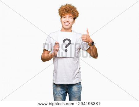 Young handsome man holding paper with question mark happy with big smile doing ok sign, thumb up with fingers, excellent sign
