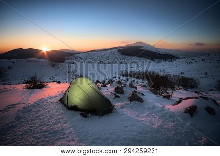 illuminated tent on the snow of winter landscape in Nebrodi Park at the sunrise, on background frozen Trearie Lake and Etna Volcano, Sicily  