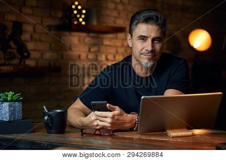 Older white man sitting at desk in dark room at home, working, using phone and tablet