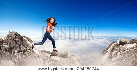Redhead Woman Jumps Through The Gap Between Hills Over Cliff On Blue Sky Background.