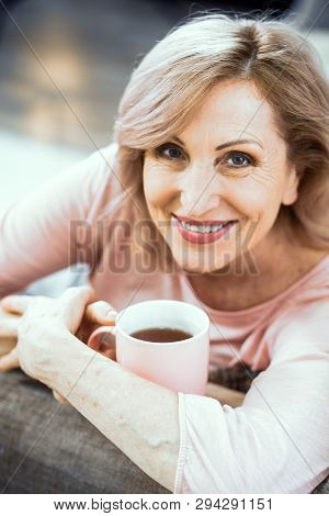 Smiling Woman Looks Over 50 Years Old. She Is Resting In Her Living Room On The Couch. In The Hands 