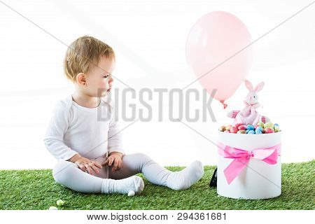 Cute Baby Sitting On Green Grass And Looking At Box With Estern Eggs, Decorative Rabbit And Air Ball