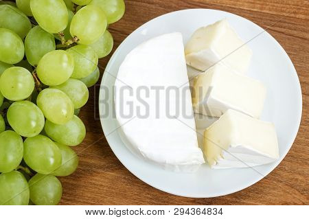 White Plate With Pieces Of Tasty Cheese Camembert And Sweet Green Grapes On A Brown Woodwn Cutting B