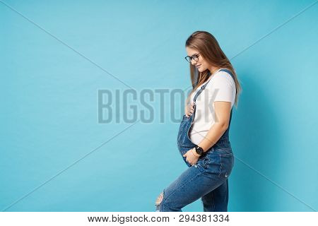 Pregnant Woman Caressing Her Belly Over Blue Background. A Cute Young Woman Wearing Glasses, And Den