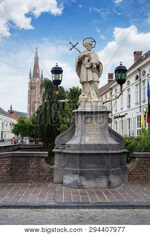 Bruges, Belgium - August 16, 2013: Scenic cityscape with statue of Johannes Nepomucenus, church tower against the blue sky in Bruges