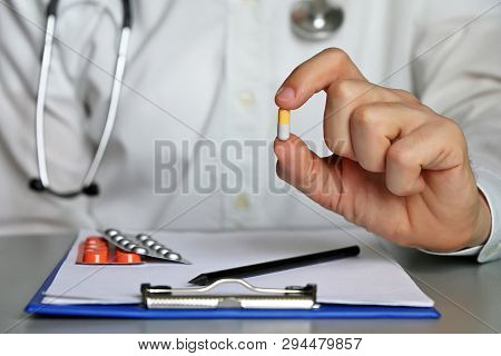 Doctor Sitting In Hospital Office With Pill In Hand, Physician At Work Giving Medication In Capsule.