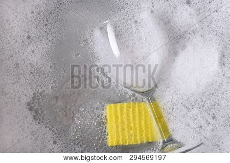 High angle view of a dishpan with a single wine glass and yellow sponge immersed in soapy water. 