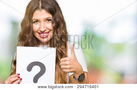 Young adult woman holding paper with question mark happy with big smile doing ok sign, thumb up with fingers, excellent sign