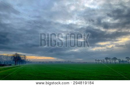 Dramatic And Colorful Sunrise Or Sunset Sky Over A Grassy Green Farmfield Lined By Trees