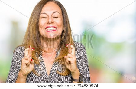 Beautiful middle age business woman smiling crossing fingers with hope and eyes closed. Luck and superstitious concept.