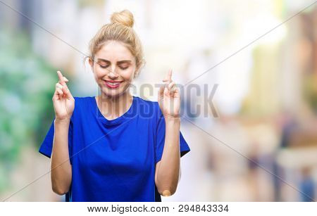 Young beautiful blonde and blue eyes woman wearing blue t-shirt over isolated background smiling crossing fingers with hope and eyes closed. Luck and superstitious concept.
