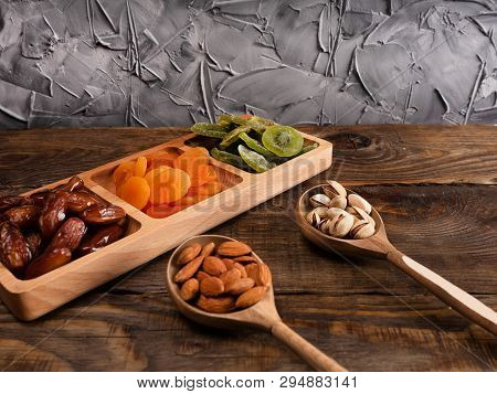 Dates, Dried Apricots And Kiwis In A Compartmental Dish And Nuts In A Wooden Spoon On A Dark Wooden 