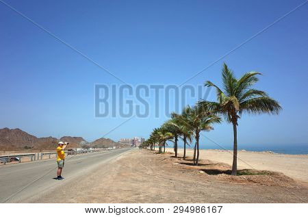 Tourist standing on road and taking photo of palm trees and sandy beach of Gulf of Oman in Fujairah, United Arab Emirates