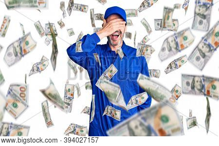 Middle age handsome man wearing mechanic uniform smiling and laughing with hand on face covering eyes for surprise. blind concept.