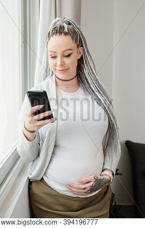 Smiling Attarctive Young Pregnant Woman With Dreadlocks Standing At Window, Touching Belly And Readi