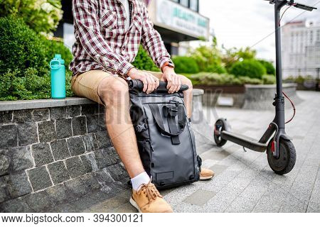 Young Caucasian Man Opens A Black Roll Top Backpack While Sitting On A Bench Near An Electric Scoote