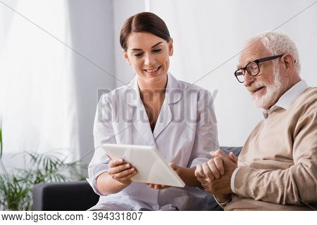 Social Worker Showing Dgital Tablet To Senior Man Sitting With Clenched Hands