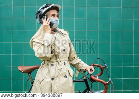 Woman in protective maskin, in helmet using her smartphone device. Female go to work on his sport retro bike. Girl standing on green background.