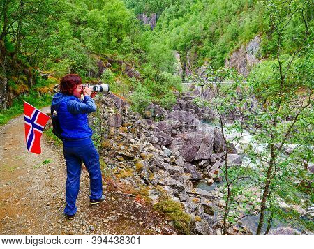 Tourist Woman With Norwegian Flag And Camera Walking Trough Mountains Path In Allmannajuvet Area Sau