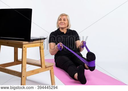 An Older Woman Sits On A Mat In Front Of A Computer And Does Exercises With An Elastic Band. Pandemi