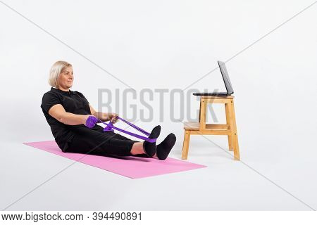 Senior Woman Sitting On A Fitness Mat In Front Of The Computer And Doing Exercises With An Elastic B