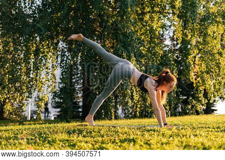 Side View Of Flexible Young Woman Practicing Yoga In Down Facing Dog Pose On One Leg, City Park At S
