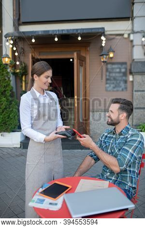 Bearded Male Paying Bill With Smartphone In Open Air Cafe, Waitress Holding Payment Terminal