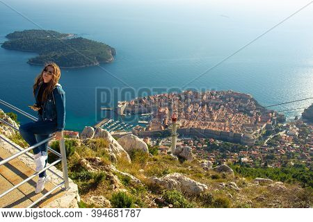Attractive Brunette Traveller Sitting On A Railing Above The City Of Dubrovnik, Viewpoint On Srd Mou