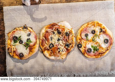 Several Ready-made Mini Pizza, Pizzetta With Sausage And Bacon, Jamon On A Linen Napkin On A Wooden 