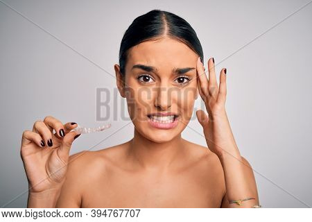 Young beautiful brunette woman holding dental aligner orthodontic to teeth corretion stressed with hand on head, shocked with shame and surprise face, angry and frustrated. Fear and upset for mistake.
