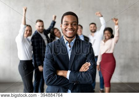 Successful Black Businessman Standing In Front Of Joyful Business Team In Modern Office, Smiling To 