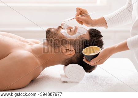 Side View Of Bearded Man Getting Face Treatment At Modern Spa Salon. Female Attendant Applying Nouri
