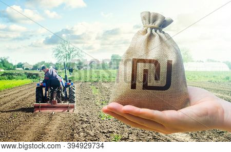 Hand Holds Out A Israeli Shekel And A Farmer On A Tractor Works The Farmer's Field. Financial Suppor