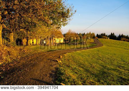 Autumn Hilly Landscape With Country Road At Sunset Time. Colorful Trees And Meadows Around Cottage U