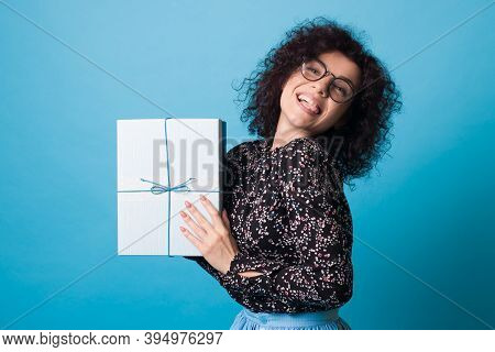 Adorable Woman With Curly Hair And Glasses Is Showing Her Tongue While Holding A Present On A Blue S