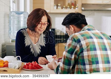 Mature Asian Woman Asking Son To Help Her With Paying Bills When Thay Are Sitting At Kitchen Table