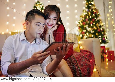 Happy Young Asian Couple Enjoying Cozy Winter Evening And Watching Christmas Movie On Screen Of Tabl