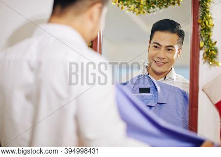Handsome Young Asian Man Choosing Shirt For Christmas Celebration And Looking At Mirror