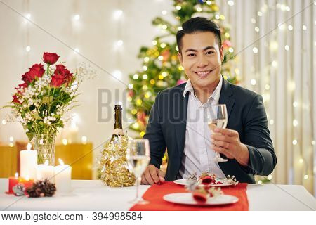 Portrait Of Happy Handsome Young Man Sitting At Christmas Dinner Table With Glass Of Champagne