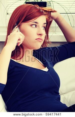 Dissapointed and upset young woman sitting and talking by the phone. During phone conversation girl is holding his head and cellphone. 