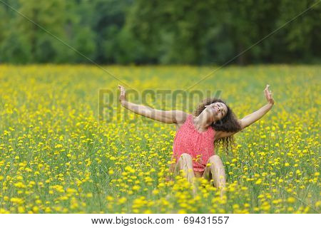 Beautiful young woman stretching her arms high in the air with a smile of pleasure and satisfaction as she sits in a rural meadow full of colorful yellow flowers, with copyspace