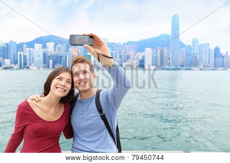 Selfie tourists couple taking self-portrait picture photos in Hong Kong enjoying sightseeing on Tsim Sha Tsui Promenade and Avenue of Stars in Victoria Harbour, Kowloon, Hong Kong. Travel concept.