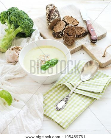 Broccoli cream soup with sunflower seeds, fresh basil and bread in bowl over white wooden table.
