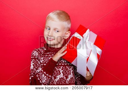 Small caucasian boy in traditional x mas knitted clothing isolated on red background excited looking at gift hold it and guess what`s inside curious dreamy can`t wait to open it