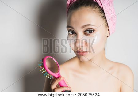A girl with a mask under her eyes looks in the mirror on the hairbrush