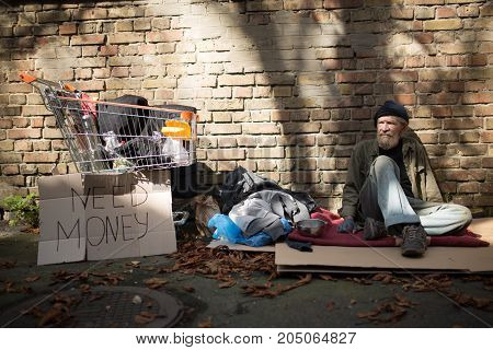 Homeles man sitting on ground in the strteet. Tramp sitting on card board by the brick wall.