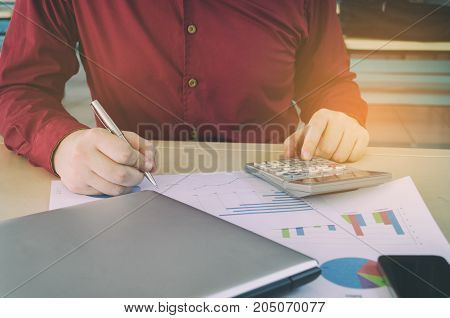 young man working calculate finance cost with calculator and writing with business strategy diagram report laptop computer smart phone on desk at home office income and expenses economy concept