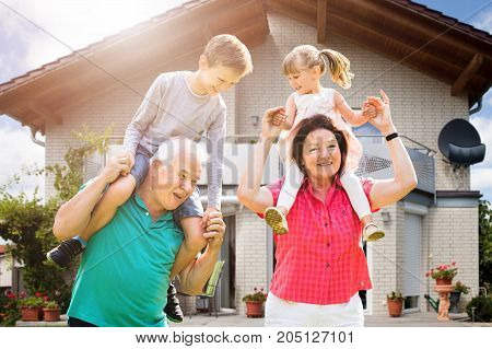 Smiling Grandchildren's Sitting On Grandparent's Shoulder Enjoying Outside Their House
